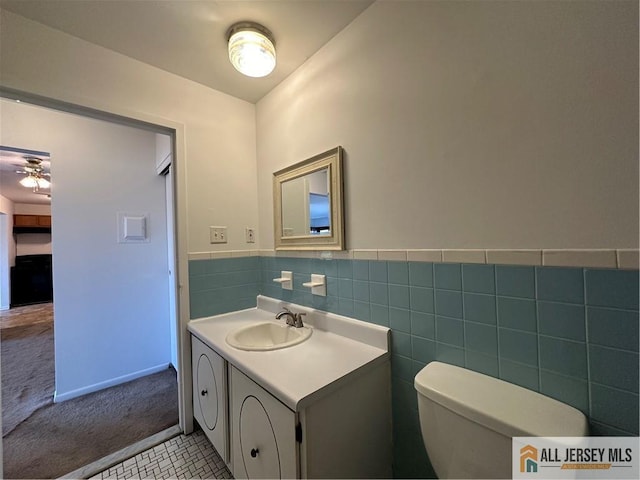 half bath with tile walls, toilet, ceiling fan, vanity, and wainscoting