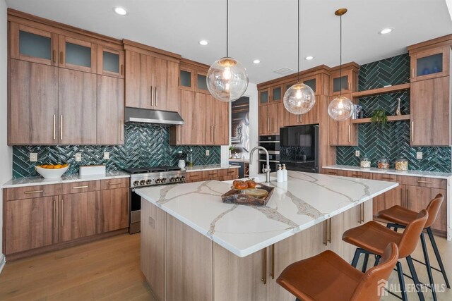 kitchen featuring light stone counters, hanging light fixtures, black refrigerator, stainless steel stove, and a kitchen island with sink