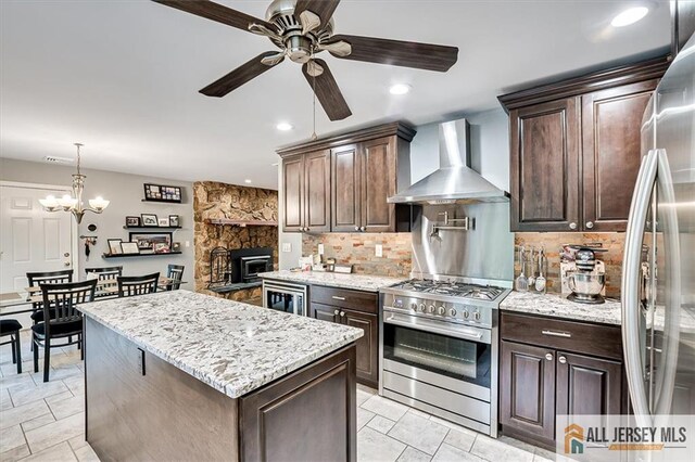 kitchen with pendant lighting, wall chimney range hood, appliances with stainless steel finishes, dark brown cabinetry, and light stone counters