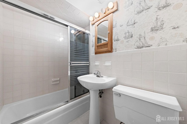 full bath featuring a wainscoted wall, toilet, shower / bath combination with glass door, tile walls, and wallpapered walls