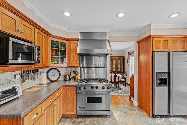 kitchen featuring appliances with stainless steel finishes, wall chimney range hood, brown cabinets, dark countertops, and ornamental molding