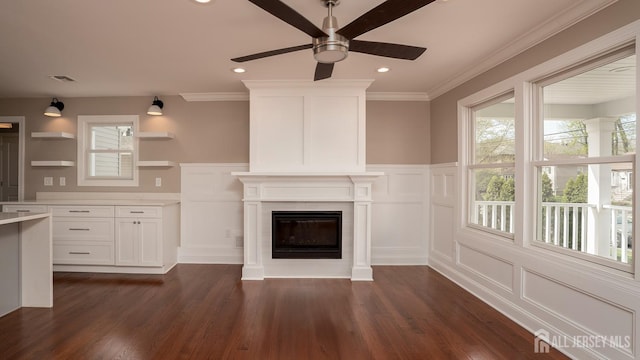 unfurnished living room with a ceiling fan, a decorative wall, dark wood-type flooring, a glass covered fireplace, and crown molding