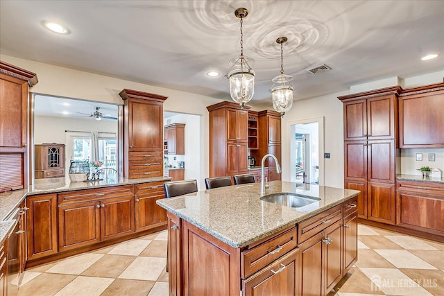 kitchen featuring visible vents, a sink, light stone counters, hanging light fixtures, and a kitchen island with sink