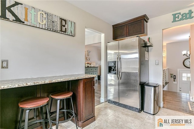 kitchen with stainless steel refrigerator with ice dispenser, dark brown cabinets, a kitchen breakfast bar, light tile patterned floors, and light stone counters