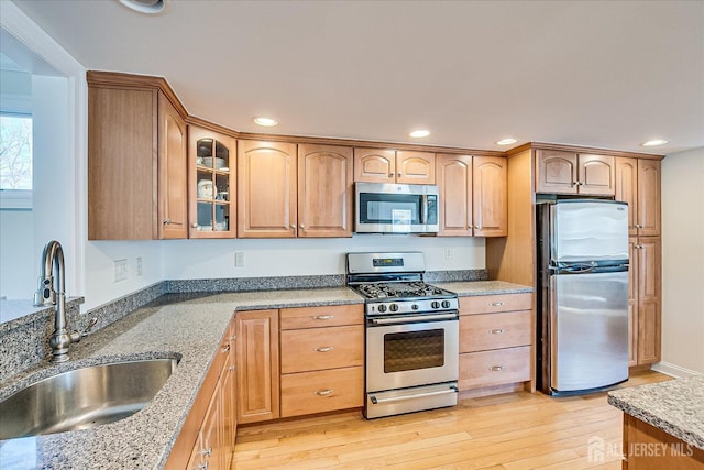 kitchen featuring light stone countertops, light wood finished floors, a sink, stainless steel appliances, and glass insert cabinets