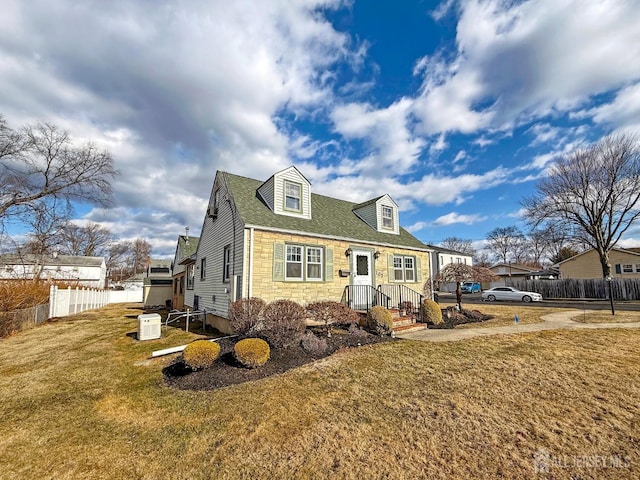 new england style home with stone siding, roof with shingles, a front yard, and fence