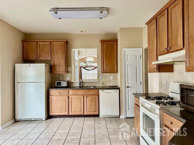kitchen featuring white appliances, under cabinet range hood, dark countertops, a sink, and brown cabinets