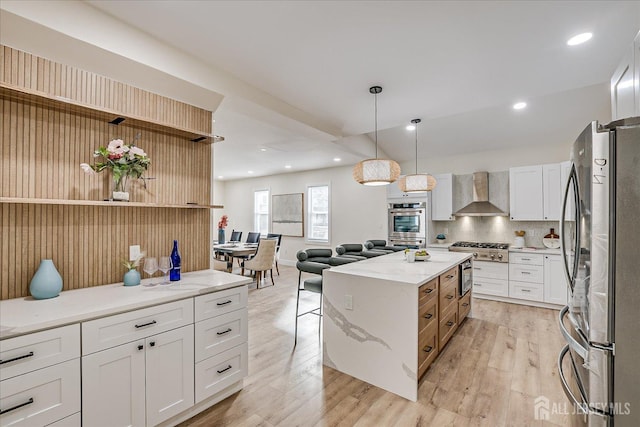 kitchen with white cabinets, appliances with stainless steel finishes, a kitchen island, decorative light fixtures, and wall chimney range hood