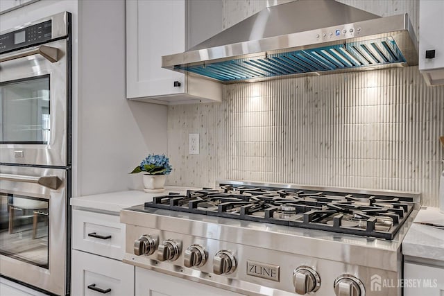 kitchen with wall chimney range hood, decorative backsplash, white cabinetry, appliances with stainless steel finishes, and light stone counters