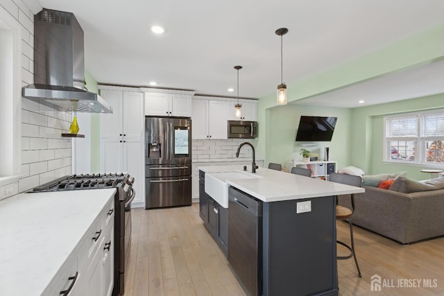 kitchen featuring appliances with stainless steel finishes, pendant lighting, white cabinets, wall chimney range hood, and a center island with sink