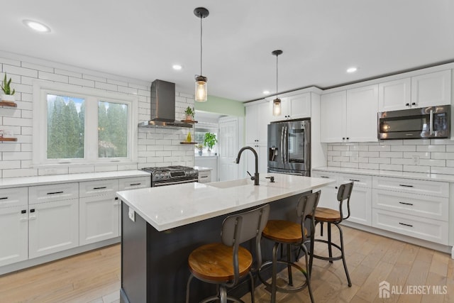 kitchen with wall chimney exhaust hood, stainless steel appliances, a center island with sink, and white cabinets