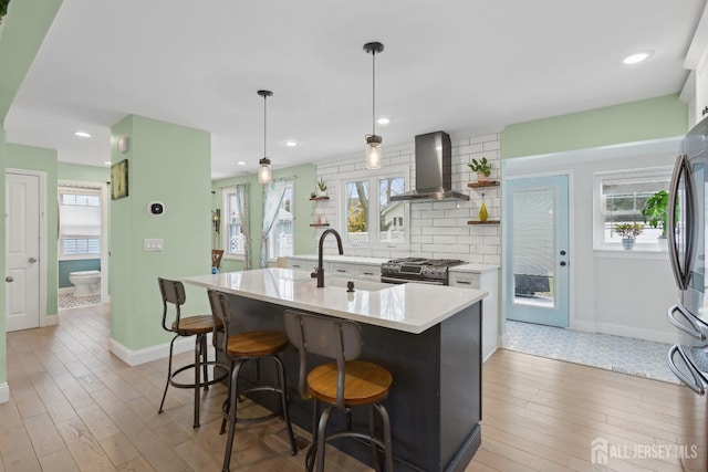 kitchen featuring open shelves, white cabinetry, wall chimney exhaust hood, an island with sink, and decorative light fixtures