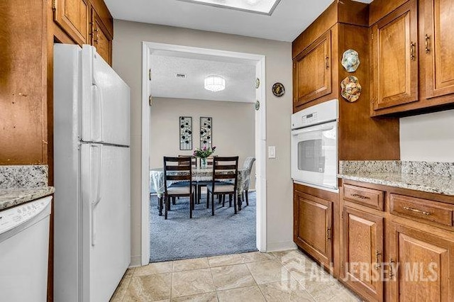 kitchen with brown cabinetry, white appliances, light carpet, and light stone counters