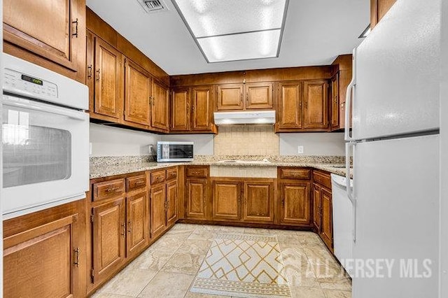 kitchen featuring white appliances, light stone counters, brown cabinetry, visible vents, and under cabinet range hood