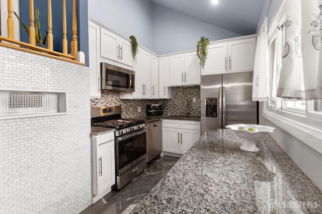 kitchen featuring stainless steel appliances, white cabinetry, vaulted ceiling, decorative backsplash, and dark stone counters