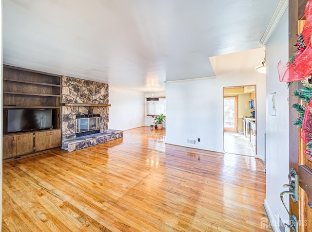 living room with built in shelves, a stone fireplace, crown molding, and light hardwood / wood-style flooring