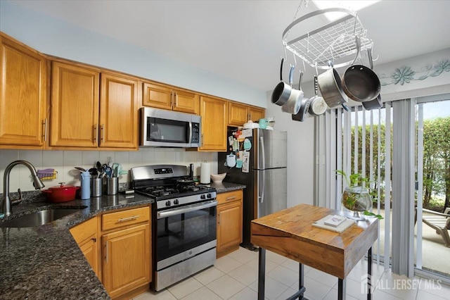 kitchen featuring brown cabinetry, appliances with stainless steel finishes, a sink, light tile patterned floors, and dark stone counters