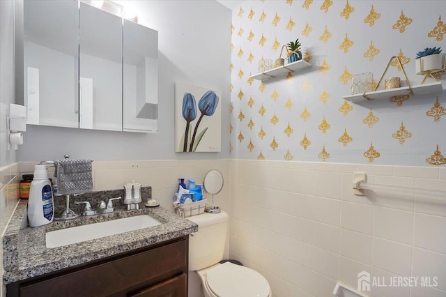 bathroom with wainscoting, wallpapered walls, toilet, vanity, and tile walls
