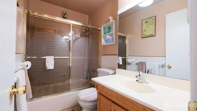 bathroom featuring toilet, vanity, bath / shower combo with glass door, and a wainscoted wall