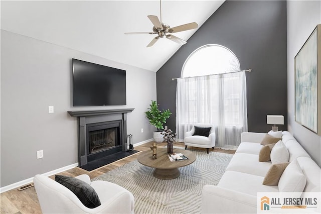 living area with baseboards, a ceiling fan, a fireplace with flush hearth, wood finished floors, and high vaulted ceiling