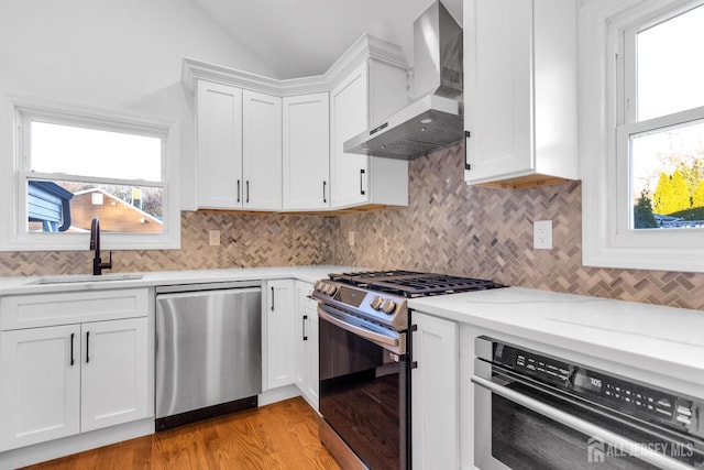 kitchen featuring backsplash, appliances with stainless steel finishes, white cabinets, a sink, and wall chimney exhaust hood