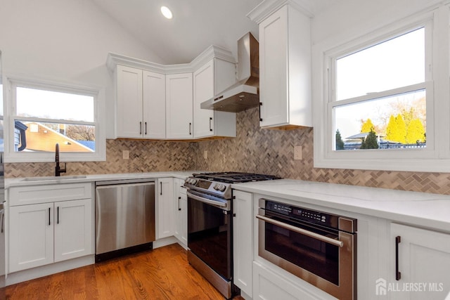 kitchen with stainless steel appliances, lofted ceiling, white cabinetry, a sink, and wall chimney range hood