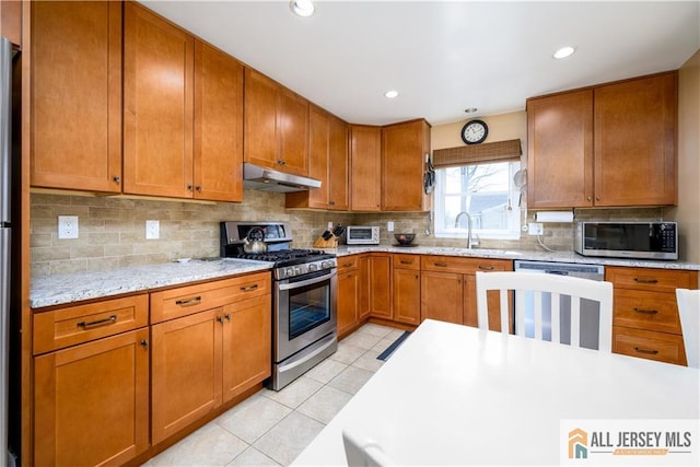 kitchen featuring light tile patterned floors, brown cabinetry, a sink, under cabinet range hood, and appliances with stainless steel finishes