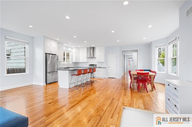 kitchen with freestanding refrigerator, wall chimney range hood, a kitchen bar, white cabinets, and recessed lighting
