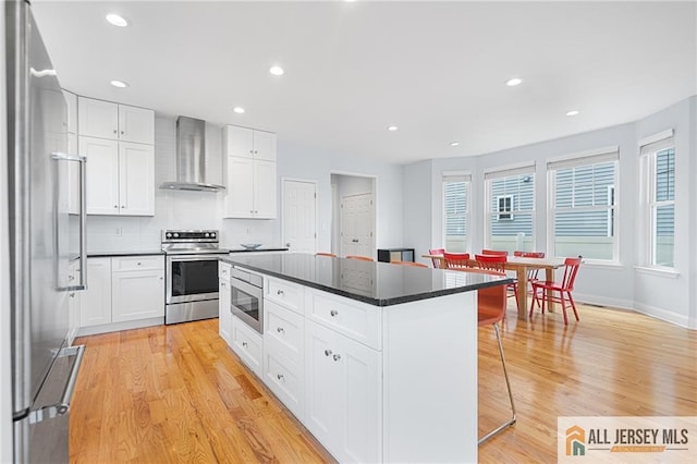 kitchen featuring wall chimney exhaust hood, stainless steel appliances, a center island, a kitchen breakfast bar, and light wood finished floors