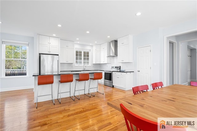 kitchen featuring stainless steel appliances, wall chimney range hood, a kitchen island, light wood-style floors, and a breakfast bar