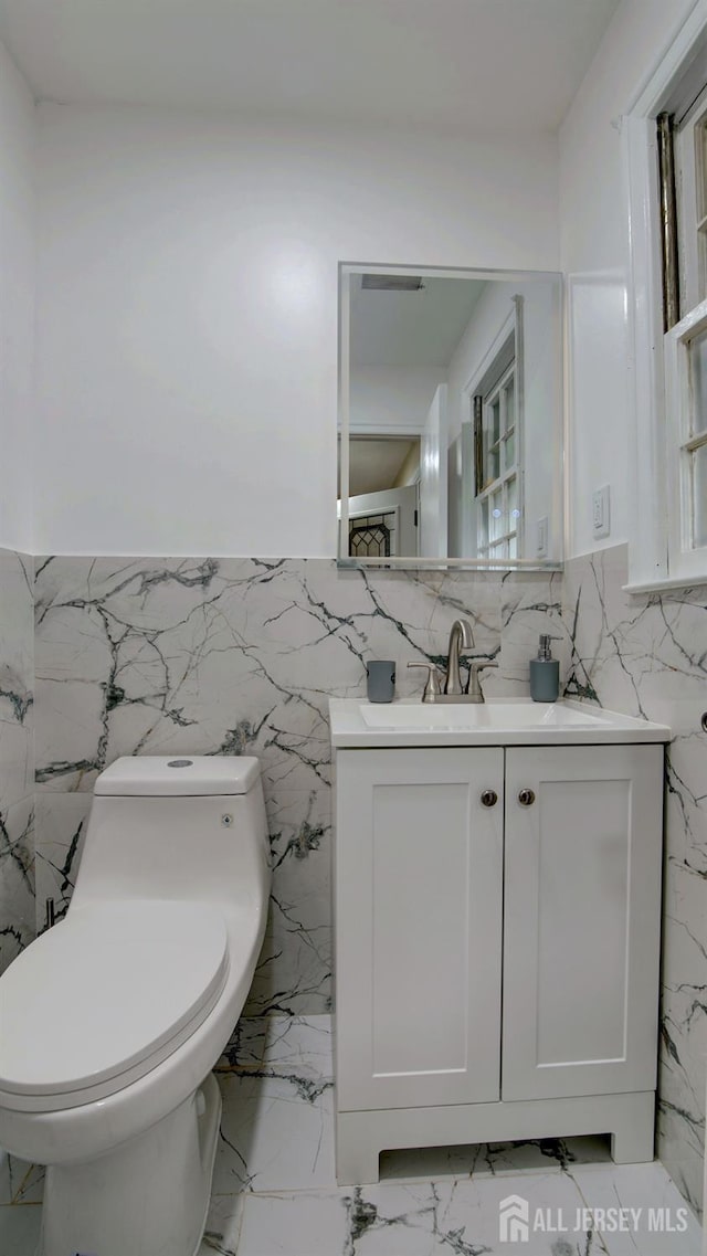 bathroom featuring marble tiled flooring, toilet, vanity, tile walls, and a wainscoted wall