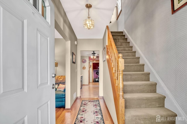 foyer entrance featuring ceiling fan with notable chandelier, stairs, wallpapered walls, baseboards, and light wood-type flooring