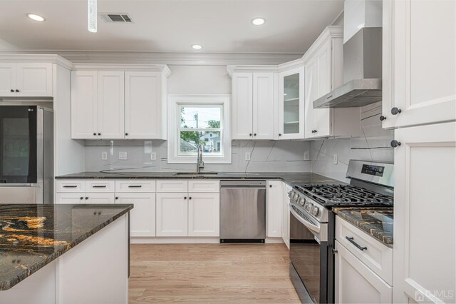 kitchen featuring wall chimney range hood, light wood-type flooring, dark stone counters, stainless steel appliances, and white cabinets