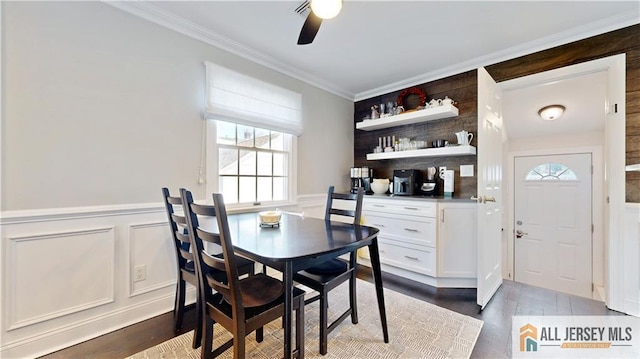 dining area with a wainscoted wall, dark wood-type flooring, ornamental molding, a decorative wall, and ceiling fan