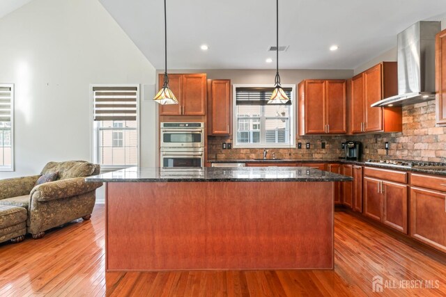 kitchen featuring pendant lighting, wall chimney range hood, appliances with stainless steel finishes, a center island, and dark stone counters