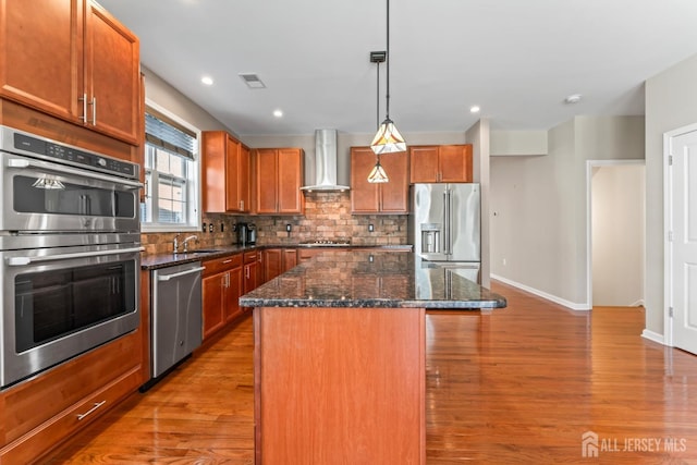 kitchen with hanging light fixtures, dark stone countertops, a kitchen island, stainless steel appliances, and wall chimney range hood