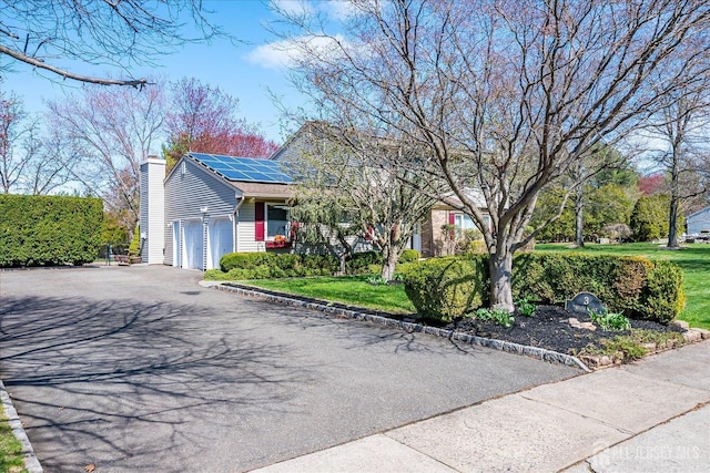 view of front of house featuring aphalt driveway, a front yard, solar panels, an attached garage, and a chimney