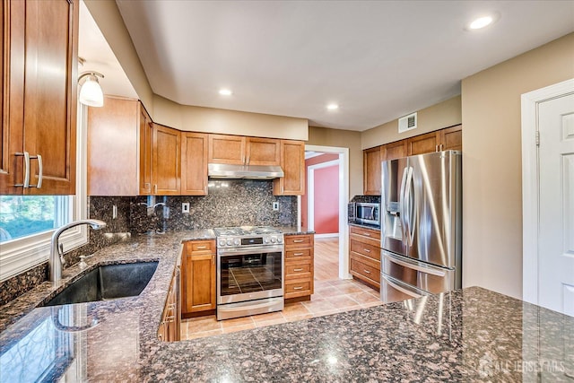 kitchen featuring dark stone counters, under cabinet range hood, a sink, stainless steel appliances, and decorative backsplash