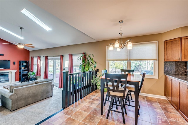 dining room with ceiling fan with notable chandelier, a glass covered fireplace, tile patterned flooring, vaulted ceiling, and baseboards