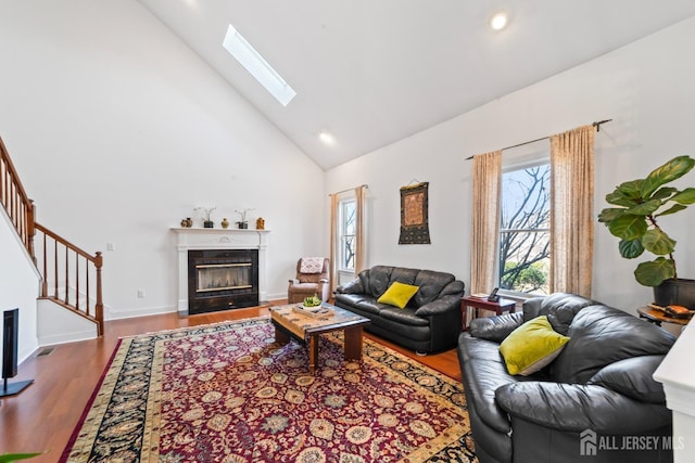 living area featuring high vaulted ceiling, a skylight, wood finished floors, a fireplace with flush hearth, and stairway