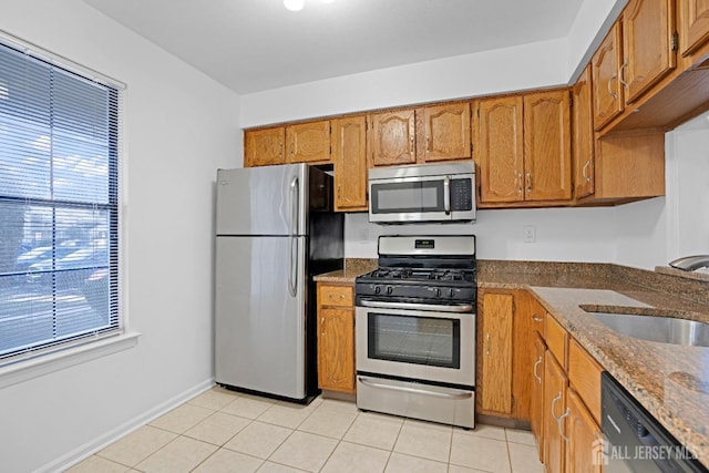 kitchen featuring light tile patterned floors, stainless steel appliances, a sink, brown cabinets, and dark stone countertops