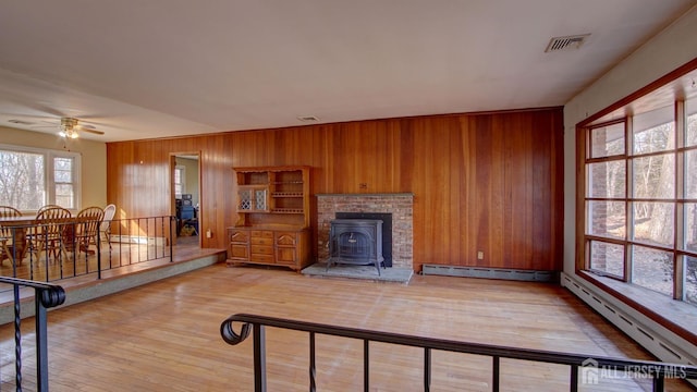 living area featuring light wood finished floors, ceiling fan, wood walls, baseboard heating, and a wood stove