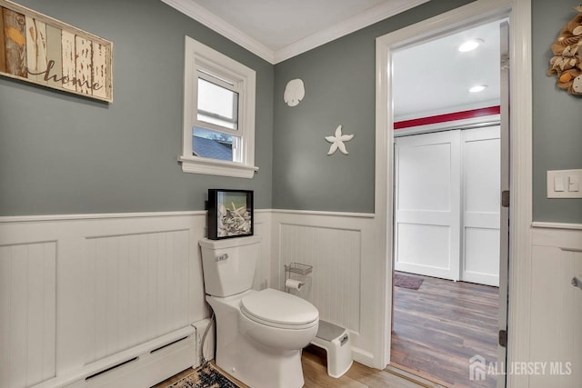 bathroom with a baseboard heating unit, wainscoting, wood finished floors, crown molding, and toilet
