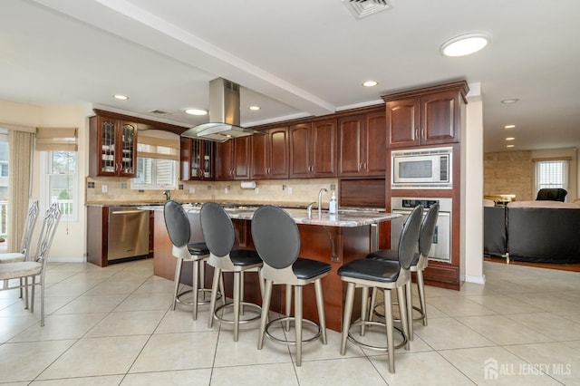 kitchen with appliances with stainless steel finishes, island exhaust hood, backsplash, glass insert cabinets, and a kitchen island with sink