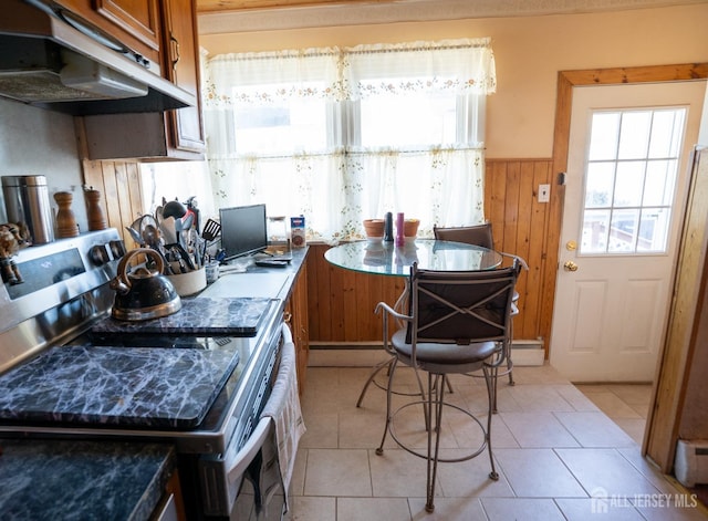 kitchen featuring brown cabinetry, a wainscoted wall, stainless steel electric range oven, under cabinet range hood, and light tile patterned flooring