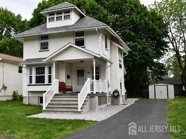 american foursquare style home with a storage unit, covered porch, asphalt driveway, an outbuilding, and a chimney