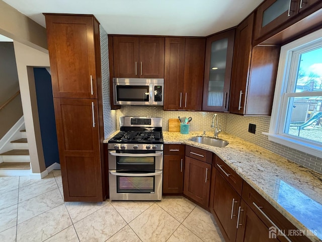 kitchen featuring a sink, light stone counters, backsplash, stainless steel appliances, and glass insert cabinets