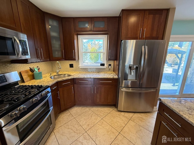 kitchen with light stone countertops, a sink, glass insert cabinets, appliances with stainless steel finishes, and backsplash