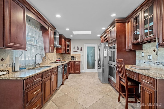 kitchen featuring stainless steel appliances, a skylight, a sink, wall chimney exhaust hood, and glass insert cabinets