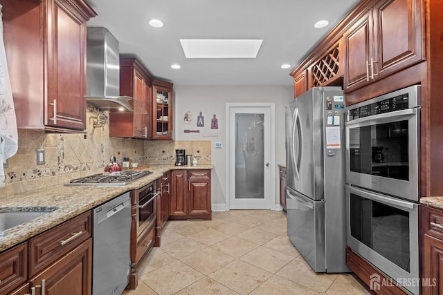 kitchen featuring light stone counters, appliances with stainless steel finishes, decorative backsplash, wall chimney exhaust hood, and glass insert cabinets